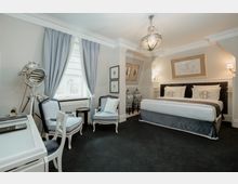 Elegantes Hotelzimmer mit hellen Wänden, dunklem Teppichboden und hochwertigen Möbeln, darunter ein großes Doppelbett, zwei Sessel und ein Schreibtisch. Die Einrichtung umfasst dekorative Elemente wie blaue Vorhänge, Bilderrahmen an der Wand sowie große verzierte Vasen.