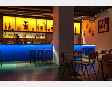 Eine stilvolle Bar mit blauer Theke und gelb beleuchteten Regalen, die mit Flaschen, Masken und Dekorationsgegenständen bestückt sind. Im Vordergrund stehen einfache Holztische und Stühle auf einem Betonboden, während an den Wänden bunte Kunstwerke und dekorative Elemente hängen.