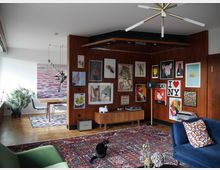 Wohnzimmer mit moderner und bunter Einrichtung, Holzwand dekoriert mit verschiedenen Kunstwerken und Postern, ein dunkelblaues Sofa, ein grüner Sessel, Teppiche mit orientalischen Mustern sowie ein Tisch- und Sitzbereich im Hintergrund. Eine schwarze Katze sitzt auf dem Teppich, und eine Zimmerpflanze steht in der Ecke.