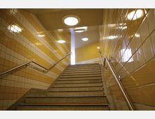 Eine Treppe in einem unterirdischen Durchgang mit gelb-weiß gefliesten Wänden. An beiden Seiten befinden sich Metallhandläufe, und runde Deckenleuchten beleuchten den Bereich.