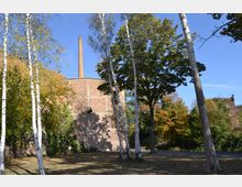 Backsteinbau mit hohem Schornstein, umgeben von Bäumen, darunter Birken und Laubbäume in herbstlichen Farben. Der Vordergrund zeigt eine Wiese mit vereinzeltem Schattenwurf, und der Himmel ist klar und blau.