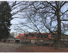 Ein zweigeschossiges Gebäude mit rotem Satteldach und mehreren Dachfenstern, teilweise von kahlen Bäumen und Ästen verdeckt, in einer ländlichen Umgebung mit offenem Gelände und Laub am Boden.