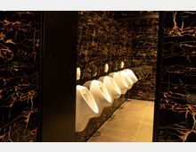 Öffentliche Herrentoilette mit einer Reihe von weißen Urinalen an einer Wand mit schwarzer Marmoroptik, durchzogen von orangefarbenen und goldenen Adern; Boden mit grauen Fliesen.