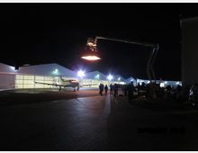 Eine nächtliche Szene auf einem Flugplatz mit einem kleinen Flugzeug vor einer Reihe beleuchteter Hangars. Im Vordergrund sind Personen und ein großer Scheinwerfer an einem Kran sichtbar, der den Bereich ausleuchtet.