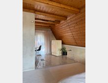 Dachgeschossraum mit Holzverkleidung an Wänden und Decke, offener Wohnbereich mit heller Einrichtung. Im Vordergrund ein cremefarbener Teppichboden, dahinter ein kleiner Essbereich mit Tisch und Stühlen sowie ein cremefarbener Retro-Kühlschrank auf einem gefliesten Boden.