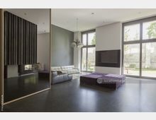 Moderner Wohnraum mit großen bodentiefen Fenstern, die Blick auf einen Garten ermöglichen. Der Raum ist mit einer weißen Ledercouch, einem violetten Ottomane sowie einem eingebauten schwarzen Flachbildfernseher ausgestattet und hat polierten dunklen Boden.