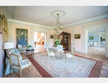 Helles, elegantes Wohnzimmer in historischem Stil mit pastellfarbenen Wänden, einem großen Teppich, filigranen Möbeln und einem Kronleuchter. Im Hintergrund sind weitere Räume mit klassischen Möbeln und Gemälden sichtbar.