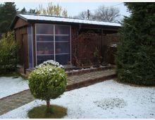 Holzschuppen in einem verschneiten Garten mit einer gepflasterten Fläche davor. Im Vordergrund ein kleiner, schneebedeckter Nadelbaum und im Hintergrund weitere Bäume.