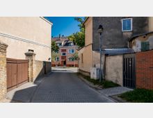 Schmale gepflasterte Gasse zwischen Gebäuden mit vergilbtem und grauem Putz, flankiert von Ziegelmauerwerk und Holztoren, führt zu einer Straße mit einem mehrfarbigen Wohnhaus und wenig Bäumen im Hintergrund.