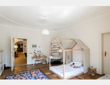 Ein helles Kinderzimmer mit Holzfußboden und einer weißen Wandfarbe. Im Raum stehen ein kleines Hausbett aus Holz, ein niedriger Kindertisch mit Stühlen, ein Teppich mit buntem Muster und ein Kachelofen. Im Hintergrund sieht man einen Durchgang zu einem weiteren Raum mit Holzmöbeln und warmem Licht.