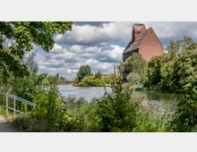 Backsteinbau überblickt einen Fluss, umgeben von dichtem Grün und Bäumen, mit einer schmalen Brücke im Hintergrund unter bewölktem Himmel. Ein Pfad mit Geländer führt am bewachsenen Ufer entlang.