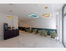 Modernes Café mit hellen Wänden und glänzendem Marmorboden. Die Einrichtung umfasst grüne gepolsterte Bänke, goldgerahmte Stühle und runde, hängende Leuchten; an der Seite steht eine Selbstbedienungs-Kaffeemaschine.