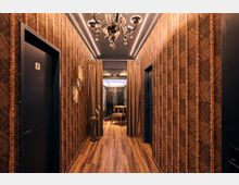 Narrow interior hallway with patterned brown wallpaper, dark doors on both sides, and wood flooring, lit by an ornate chandelier and ceiling strip lights. At the end, curtains frame an opening with a small chair, a sunburst wall mirror, and a decorative bird statue.