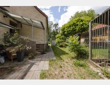 Schmaler Hof neben einem hellen Backsteinhaus mit Glasvordach über dem Eingang und einem gepflasterten Weg, der zu einem kleinen Schuppen am Ende führt. Rechts begrenzt ein Metallgitterzaun den Bereich, dahinter stehen dichte Sträucher und ein Nebengebäude mit rotem Ziegeldach.