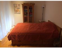 Schlafzimmer mit einem Holzbett, bedeckt von einer roten Decke, einem dekorativen Holzschrank mit Glastüren voller Bücher und Spielen, einer Stehlampe und einem kleinen Nachttisch. Links sind helle Vorhänge vor einem Fenster sichtbar.