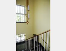 Treppenhaus mit hellgelben Wänden, großen Fenstern mit weißen Rahmen und einem goldenen Kronleuchter mit hängenden Zylinderschirmen. Das Geländer ist aus weißen Stäben und einem dunklen Holzhandlauf gefertigt, der Boden besteht aus dunklem Stein.
