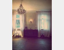 Ein schlicht eingerichteter Raum mit zwei großen Fenstern, die von weißen Vorhängen bedeckt sind. In der Mitte steht eine dunkle Kommode, darauf ein Kerzenständer und Dekorationsartikel, im Hintergrund hängt ein Kronleuchter von der Decke.