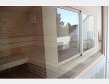 Innenansicht einer Sauna mit Holzwänden und -bänken, reflektiert in einer großen Glasscheibe. Im Hintergrund sind durch das Fenster ein Raum mit einem Sofa und ein Blick nach draußen erkennbar.