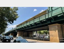Grüne Hochbahnbrücke aus Metall mit wellenförmigen Stützen und steinernen Pfeilern, die über eine belebte Straße mit wartenden Autos und Bäumen auf beiden Seiten führt. Im Hintergrund sind Gebäude und Verkehrsbereiche sichtbar.