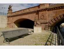 Steinbrücke mit roten Sandsteinbögen und dekorativer Verzierung, flankiert von Statuen, darunter ein Fabelwesen und ein Löwe; im Vordergrund eine Holzbank auf einem Pflasterweg, daneben ein Geländer entlang eines Kanals.