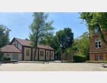 Ein kleiner, gepflasterter Platz mit einem einstöckigen Gebäude mit roten Dachziegeln und bogenförmigen Fenstern links, einem Sportbereich mit Zaun im Hintergrund und einem teilweise sichtbaren, mehrstöckigen Backsteingebäude rechts. Im Umfeld stehen mehrere Bäume und Bänke.