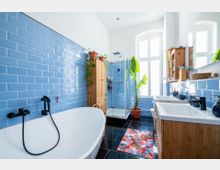 Badezimmer mit Wänden aus blauen Fliesen, einer freistehenden Badewanne mit schwarzer Armatur, einer Glasdusche und großen weißen Fenstern. Es gibt Holzmöbel, Zimmerpflanzen und eine farbige Badematte auf einem schwarzen Fliesenboden.