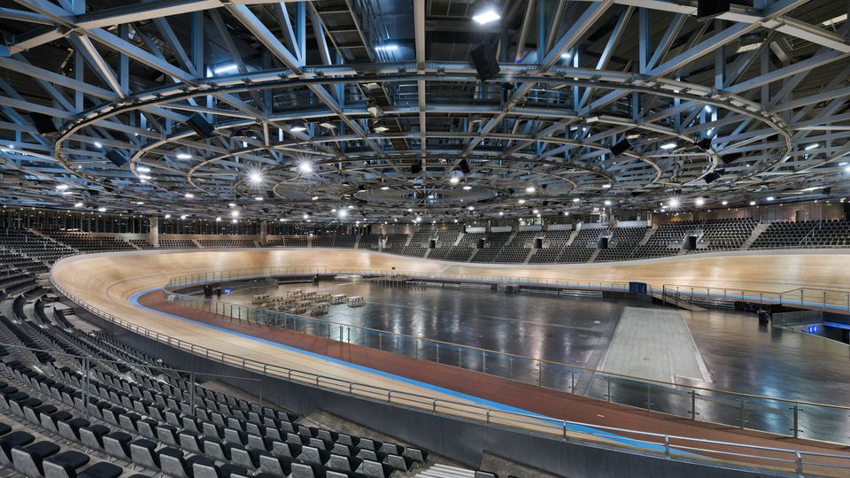 Innenansicht einer großen, modernen Radrennbahn-Halle mit ovaler Holzbahn und großem Innenfeld, umgeben von abgestuften Tribünen mit schwarzen Sitzreihen. Über der Arena spannt sich eine weitläufige Stahlträger-Decke mit integrierter Beleuchtung und hängender Veranstaltungstechnik.