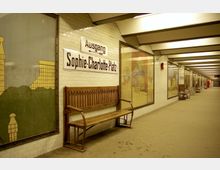 U-Bahn-Station mit der Beschilderung "Ausgang Sophie-Charlotte-Platz". Die Wände sind mit weißen Fliesen verkleidet und dekoriert mit großflächigen Illustrationen; im Vordergrund steht eine hölzerne Sitzbank.