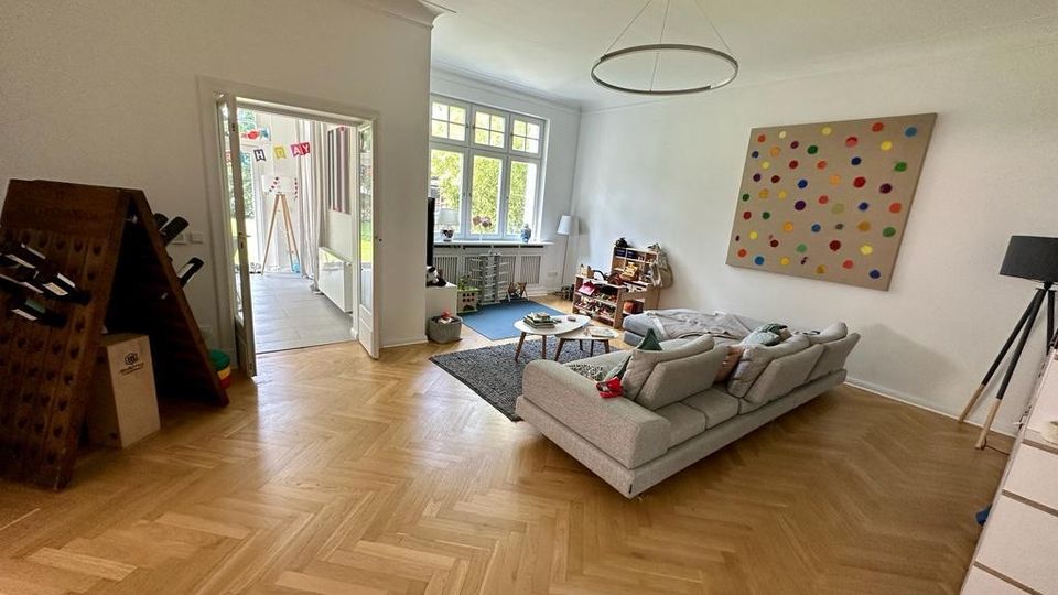 Wohnzimmer mit hellem Fischgrätenparkett, einer großen grauen Sofalandschaft und einem bunten Wandbild. Im Hintergrund befindet sich eine Tür, die zu einem Bereich mit Fliesenboden und großen Fenstern führt.