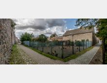 Ein ländlicher Weg mit Kopfsteinpflaster und Gehweg führt an einer Steinmauer und einem Grundstück mit Drahtzaun vorbei, hinter dem Hühner gehalten werden. Im Hintergrund stehen mehrere einfache Gebäude mit Ziegeldächern, umgeben von Bäumen, unter einem bewölkten Himmel.