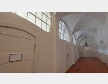 Ein heller Flur in einem historischen Gebäude mit weißen Wänden und einer gewölbten Decke. Entlang des Korridors befinden sich Holztüren mit Beschilderungen, große Fenster mit Sprossen lassen Tageslicht herein, und eine Hängelampe ist in der Mitte befestigt.