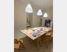 Heller Atelierraum mit großem Holzarbeitstisch und mehreren Holzstühlen, auf dem Tisch liegen Papier, Scheren und Bastelmaterialien. Von der hohen Decke hängen drei weiße Pendelleuchten; im Hintergrund stehen eine kleine Ablage und ein großes Bild an der Wand.