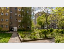 Mehrstöckige Wohnhäuser mit braunen Fassaden und weißen Fenstern, umgeben von einem gepflegten Gartenbereich mit Rasen, Bäumen und Sträuchern. Ein schmaler Gehweg führt zum Eingang eines Gebäudes, daneben ist ein Fahrrad abgestellt.