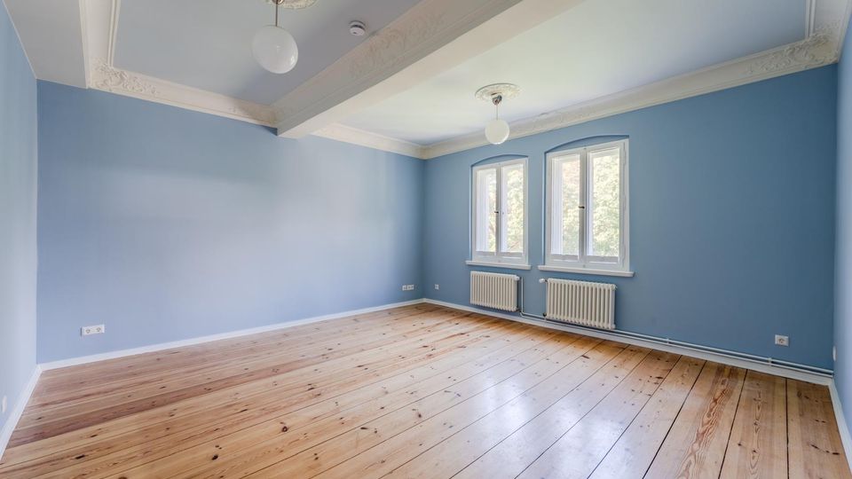 Leerer Raum mit hellblauen Wänden, breiten Holzdielen und hoher Decke mit Stuckleisten und zwei runden Deckenleuchten. Zwei hohe Fenster an der Stirnwand lassen Tageslicht herein; darunter stehen weiße Heizkörper.