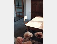 Ein minimalistischer Innenraum mit einem Holztisch und einem einfachen Stuhl auf einem Betonboden. Im Hintergrund sind eine Holzwand mit Schranktüren und eine Glastür mit Holzrahmen zu sehen; im Vordergrund unscharfe Blumen in einer Vase.