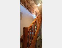 Holztreppe in einem Innenraum mit geländerartigen Holzstreben, Teppichstufen und einer Decke aus Holzpanelen. Rechts unten ist ein massiver Tresor zu sehen.