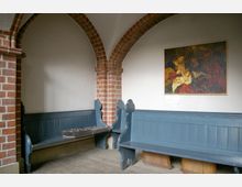Ecke eines Kircheninnenraums mit weißen Wänden, roten Backsteinbögen und blauen Holzbänken. An der Wand hängt ein Gemälde mit religiösem Motiv.