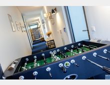 Ein Tischkicker steht in einem hellen Korridor mit blauem Teppichboden. An den Wänden hängen Bilder und kleine Dekorationen, während an einer Seite ein Konsolentisch mit Vasen und getrockneten Blumen steht.