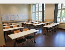 Klassenzimmer mit mehreren weißen Tischen und Holzstühlen, angeordnet in Reihen. An den Wänden hängen großformatige beschriebene Plakate, und große Fenster mit Gitterrahmen lassen Tageslicht herein.