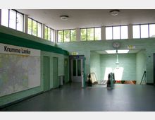 U-Bahn-Station mit hellgrünen Wandfliesen, großen Fenstern, einer Wandkarte der Umgebung mit der Aufschrift "Krumme Lanke", einer Rolltreppe, einem Fahrstuhl und einer analogen Uhr über einem Durchgang.