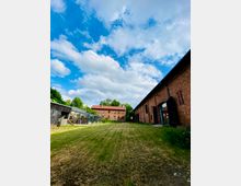 Weitläufiger Hof mit gemähtem Rasen, umgeben von roten Backsteinbauten, darunter ein großes Gebäude rechts mit offenen Toren und ein kleineres Haus im Hintergrund mit roten Dachziegeln. Links befinden sich Glasanbauten und ein eingezäunter Bereich, im Hintergrund stehen Bäume unter einem bewölkten Himmel.