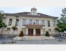 Eingangsbereich einer Bibliothek in einem historischen Gebäude mit hellem Putz, großen Sprossenfenstern und einem vorstehenden Vordach. Links und rechts des gepflasterten Vorplatzes befinden sich bepflanzte Blumenkübel und Straßenlaternen.