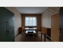 Kleines Büro mit schlichter Einrichtung aus Holz. Im Raum stehen ein Schreibtisch mit einem Stuhl davor, mehrere Holzschränke an den Wänden sowie ein Fenster mit einfacher Gardine im Hintergrund.