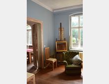 Zimmer mit Holzboden und blauen Wänden, ausgestattet mit Holzstühlen, einem grünen Sessel mit Kissen, einem Staffelei-Rahmen sowie einem großen Fenster, durch das Licht und eine grüne Außenumgebung sichtbar sind. Links führt eine geöffnete Tür in einen angrenzenden Raum mit einem Holztisch und Stühlen.