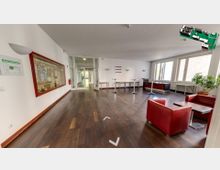 Heller Empfangsbereich mit dunklem Holzfußboden, weißen Wänden und großen Fenstern rechts, die viel Tageslicht einlassen. Links hängt eine Glastafel mit Aushängen, rechts gibt es rote Sessel und Stehtische, sowie Pflanzen in Töpfen.