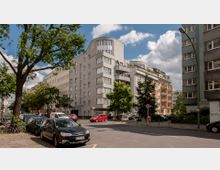 Straßenszene in einer Wohngegend mit mehrstöckigen Wohngebäuden, darunter ein modernes weißes Gebäude mit abgerundeten Ecken und Balkonen. Im Vordergrund stehen geparkte Autos und ein großer Baum, während der Himmel teils bewölkt ist.