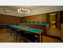 Ein historischer Gerichtssaal mit Holzwänden, einem großen Richtertisch und blauen Sitzpolstern an den Stühlen. Im Vordergrund stehen kleinere Tische und Stühle, während im Hintergrund offene Türen einen Blick in einen angrenzenden Raum mit helleren Möbeln gewähren.