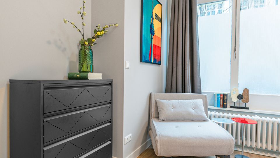 Heller Wohnraum mit hohen Decken, großem Fenster mit halbhoher Milchglasfolie und grauen Vorhängen sowie hellem Holzboden. Neben einer schwarzen Kommode mit Vase stehen eine gepolsterte Liege und ein kleiner Beistelltisch vor dem Fenster, an der Wand hängt ein farbiges Bild.