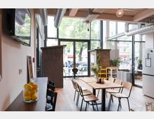 Heller Gemeinschaftsraum mit großen Glasfenstern, Holzboden und moderner Einrichtung. Ein Esstisch mit Stühlen steht im Vordergrund, während dekorative Pflanzenelemente und Küchenausstattung die Umgebung ergänzen.