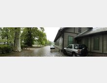 Ein gepflasterter Weg führt an einem historischen Gebäude mit Fachwerkfassade und grün gestrichenen Fensterläden vorbei. Im Vordergrund stehen zwei geparkte Autos, während im Hintergrund große Bäume und eine leicht bewaldete Landschaft zu sehen sind.